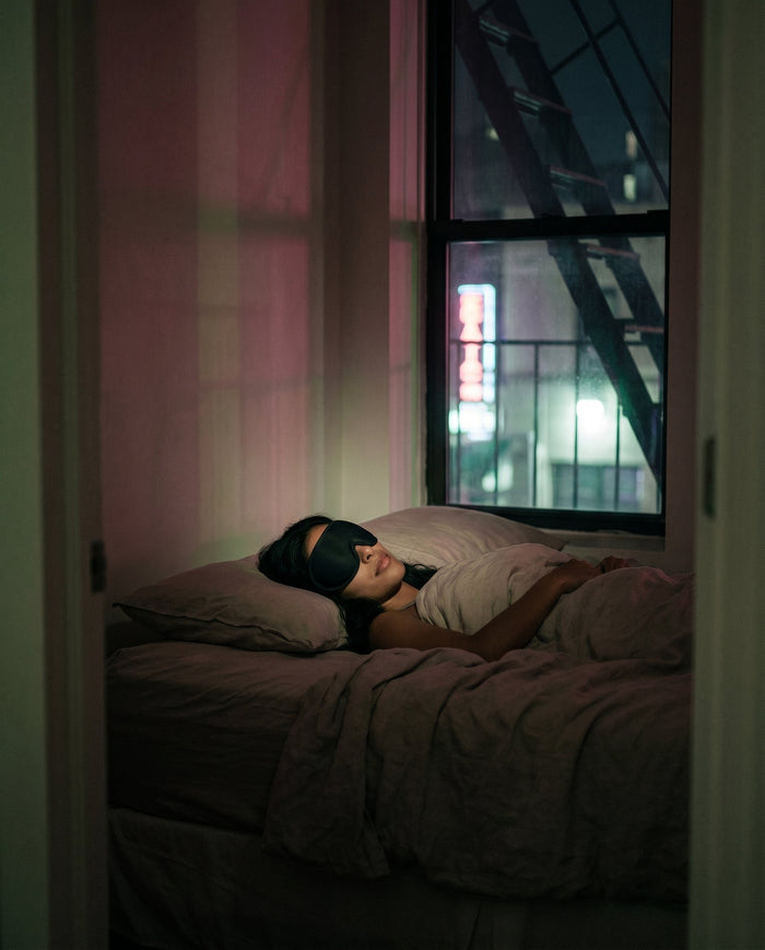 A city bedroom at night with soft linen, a warm bedside lamp and dark window light